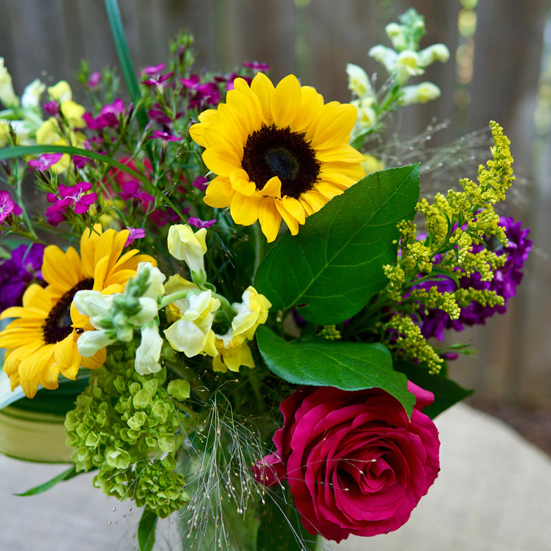 Bouquet of sunflowers, roses, and other flowers in a clear vase on a wooden surface.