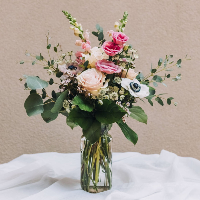 Bouquet of flowers in a clear glass vase. The bouquet is a mix of light pink and white blooms, including roses, anemones, and possibly lisianthus and snapdragons. There's also greenery, likely eucalyptus, creating a lush and romantic look. The flowers are arranged in a clear glass jar, sitting on a white fabric-covered surface against a muted beige background. The overall style is simple and elegant.