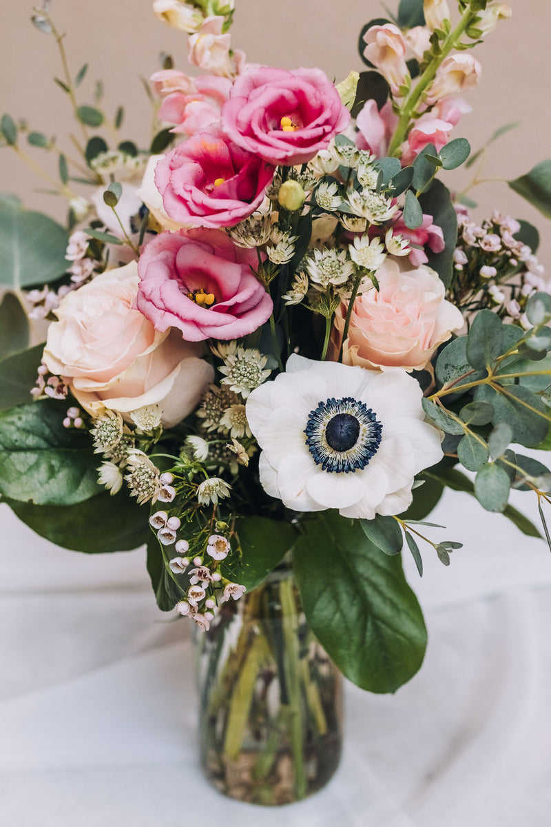 Bouquet of flowers in a clear glass vase. The bouquet is a mix of light pink and white blooms, including roses, anemones, and possibly lisianthus and snapdragons. There's also greenery, likely eucalyptus, creating a lush and romantic look. The flowers are arranged in a clear glass jar, sitting on a white fabric-covered surface against a muted beige background. The overall style is simple and elegant.