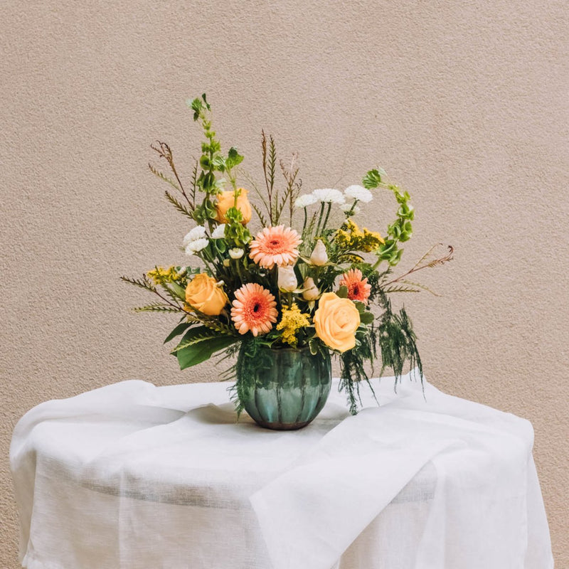 A vibrant medley of buttery yellow, rich peach, and crisp white blossoms bursts with life in a charming scalloped vase. Named for a quaint south Georgia town renowned for its Old South Day food, arts and crafts festival.