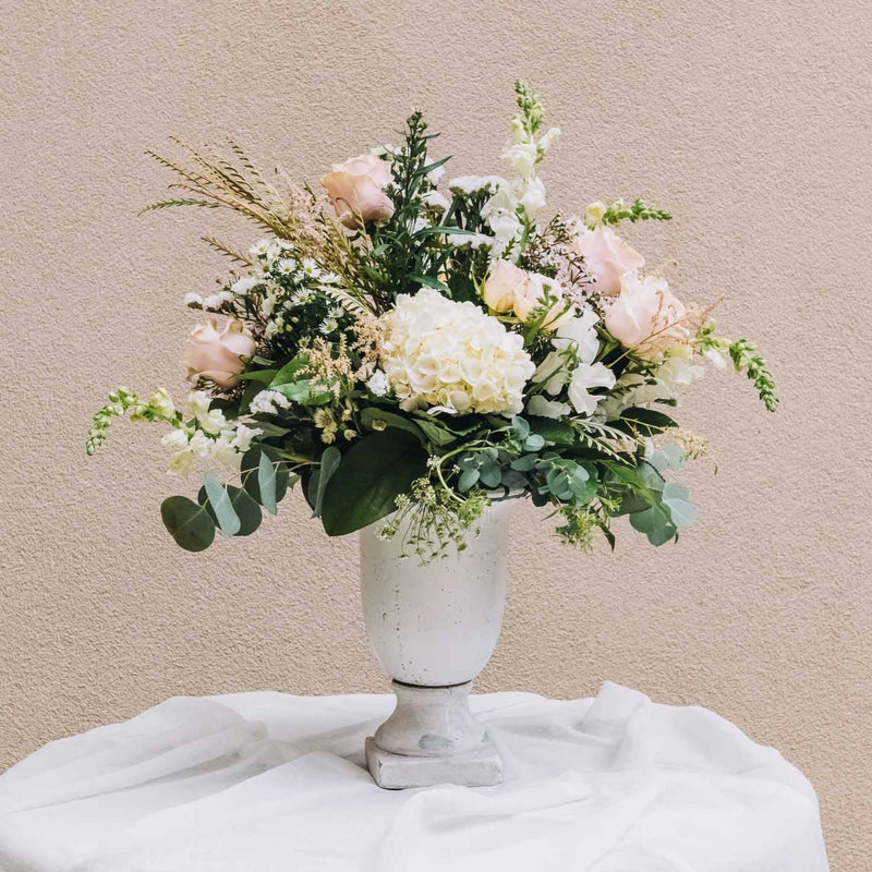 A romantic blend of soft pink and pristine white blossoms, intertwined with lush, luxurious greenery, rests gracefully in a sleek concrete urn, exuding an air of refined elegance. Inspired by a charming central Georgia city, renowned as the backdrop for beloved films like Driving Miss Daisy and Fried Green Tomatoes.