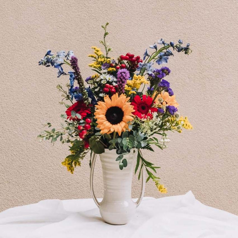Pronounced "vy-anna," this arrangement bursts with a vibrant medley of sunny yellow, fiery red, deep blue, and regal purple blooms, all artfully composed in a sleek vase that reimagines the timeless double-handled urn. Inspired by a quaint south-central Georgia city renowned for its legendary BIG PIG JIG barbecue championship.