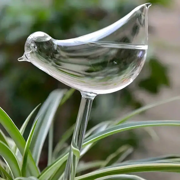 Clear glass bird-shaped watering stake nestled among green plant stems, with curved tail and curved beak spout