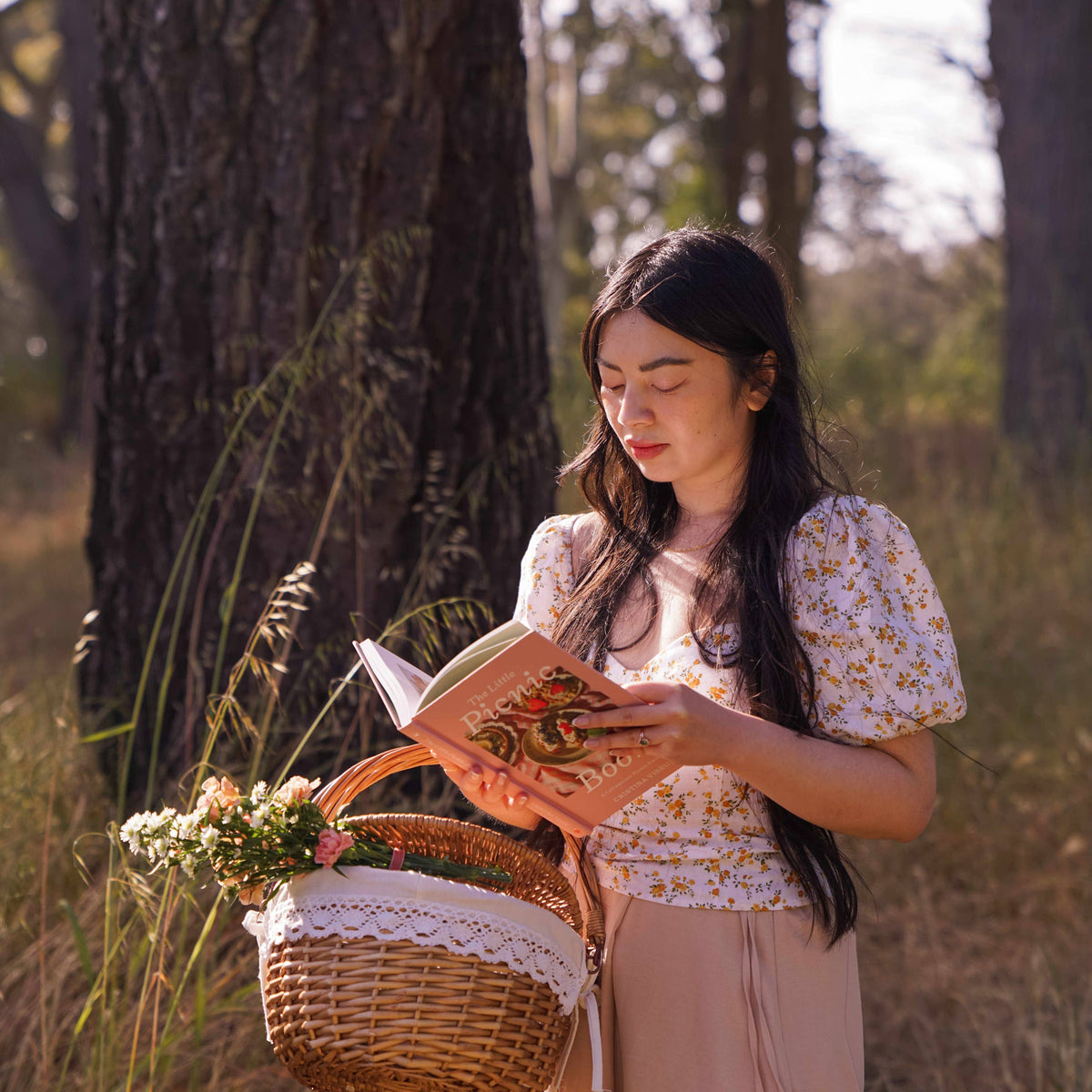 Woman in purple floral blouse reading The Little Picnic Book while holding wicker basket with flowers under trees
