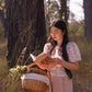 Woman in purple floral blouse reading The Little Picnic Book while holding wicker basket with flowers under trees