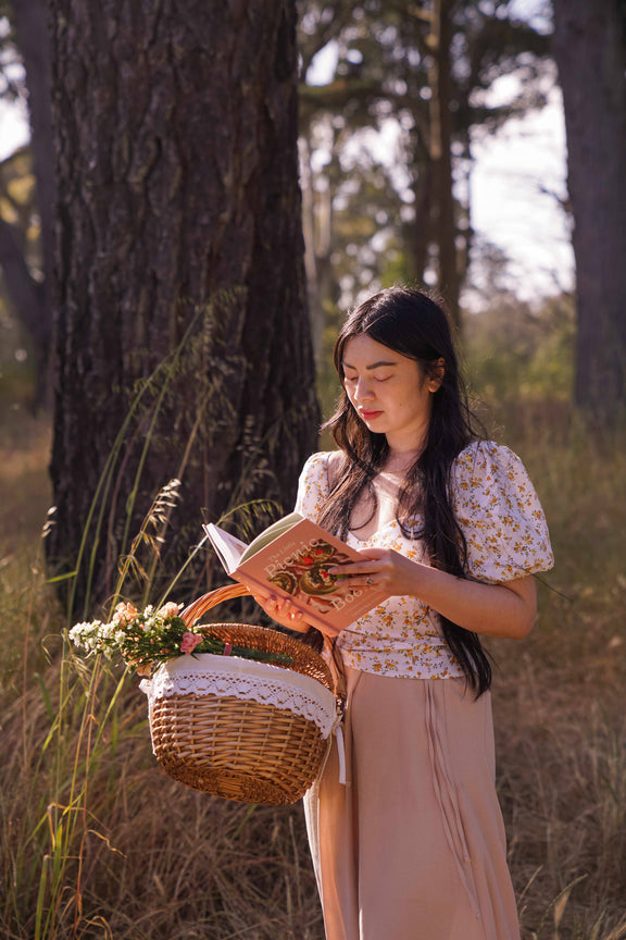 Woman in purple floral blouse reading The Little Picnic Book while holding wicker basket with flowers under trees