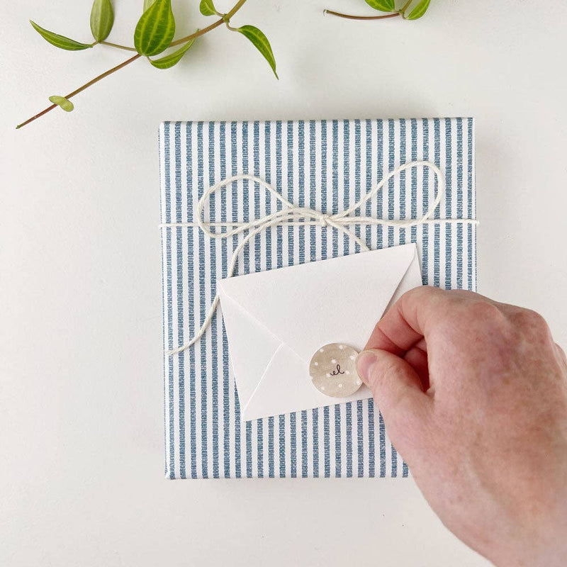 Blue and white striped mini notecard with peace sign design, wrapped twine, kraft label, and envelope, held in hand with greenery background
