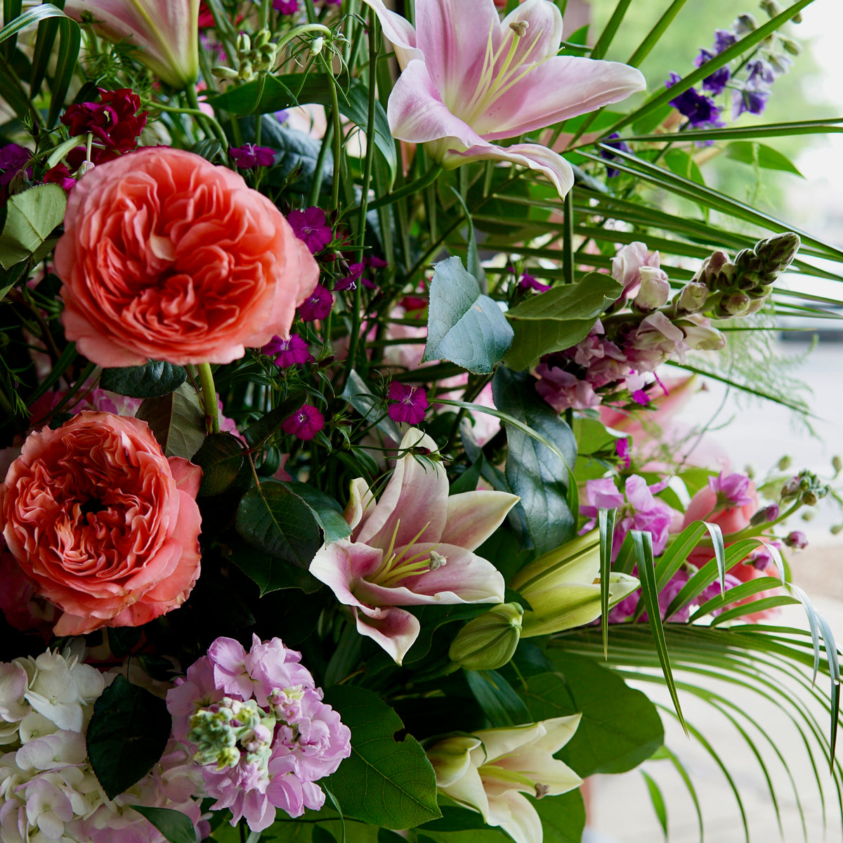 Funeral arrangement with coral garden roses, pink lilies, purple carnations and lavender, with white peonies and ornamental grass in a clear vase.