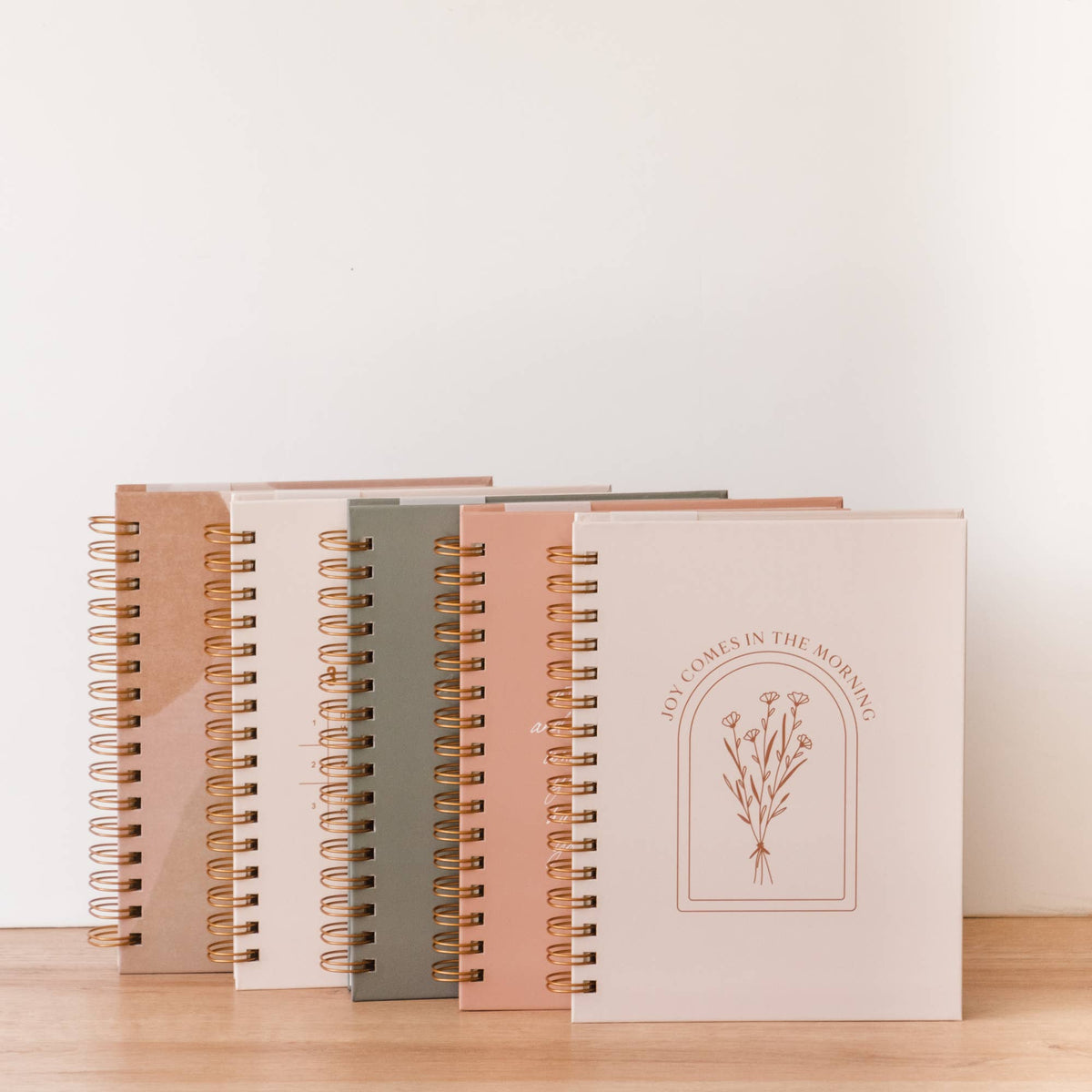 Collection of spiral-bound hardcover journals in earth tones with gold bindings; pink journal in foreground features botanical illustration and "Joy Comes in the Morning" text.