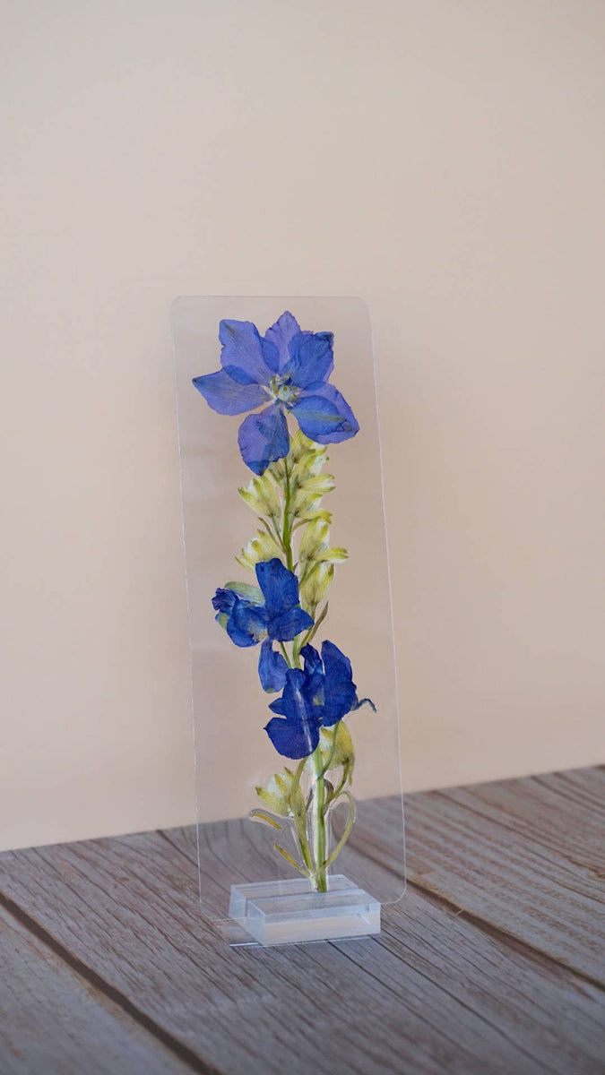 Tall rectangular transparent bookmark with blue pressed delphinium flowers, green buds and stems, white base.