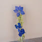 Tall rectangular transparent bookmark with blue pressed delphinium flowers, green buds and stems, white base.