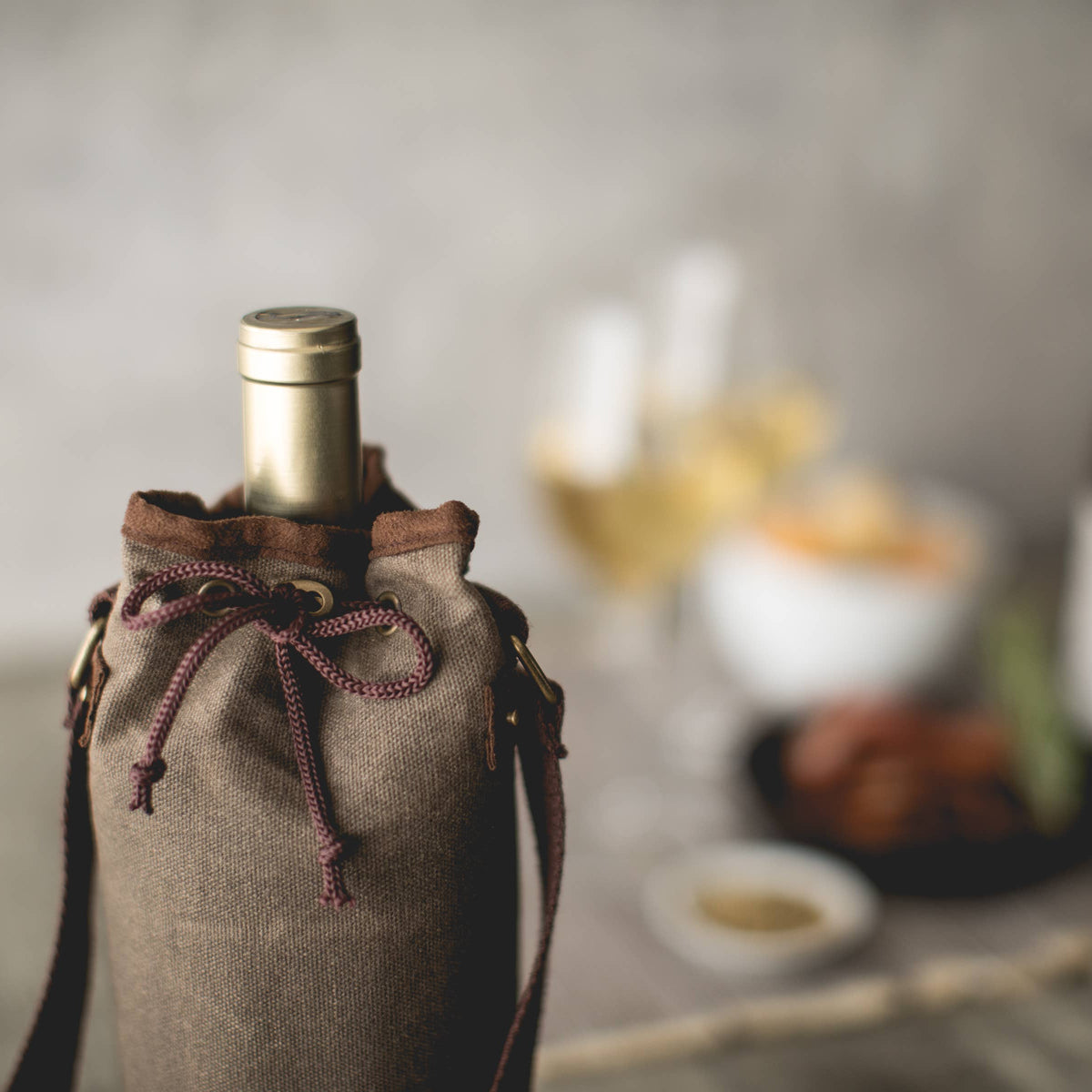 Tan waxed canvas wine bottle carrier with brown handles and burgundy drawstring, holding a wine bottle, styled with picnic spread in background