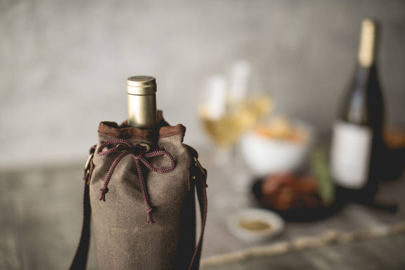 Tan waxed canvas wine bottle carrier with brown handles and burgundy drawstring, holding a wine bottle, styled with picnic spread in background