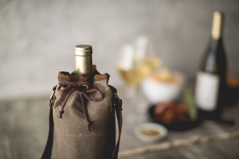 Tan waxed canvas wine bottle carrier with brown handles and burgundy drawstring, holding a wine bottle, styled with picnic spread in background