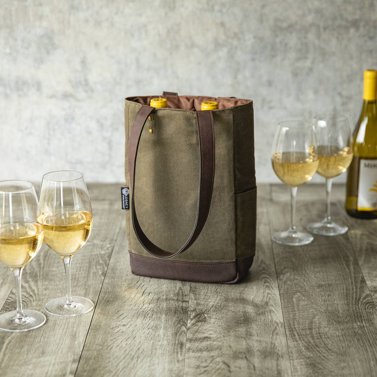 Beige canvas wine tote with brown straps holding two bottles, styled with wine glasses and a bottle on wooden surface