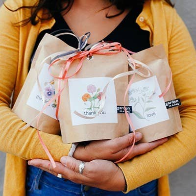 Person in mustard cardigan holding kraft paper gift bags with watercolor thank you cards, tied with coral ribbon and twine.