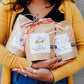 Person in mustard cardigan holding kraft paper gift bags with watercolor thank you cards, tied with coral ribbon and twine.