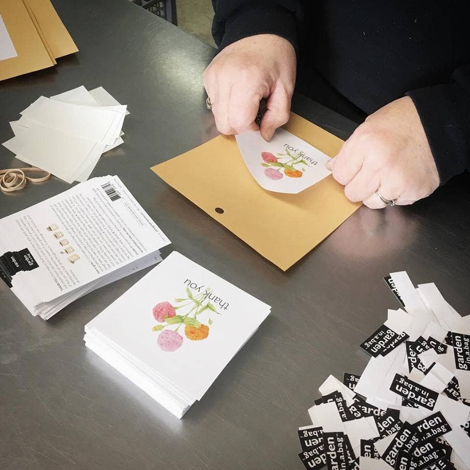 Hands opening kraft envelope with zinnia thank you cards displayed, surrounded by packaging materials including instruction cards and scattered seed packet labels on dark work surface.