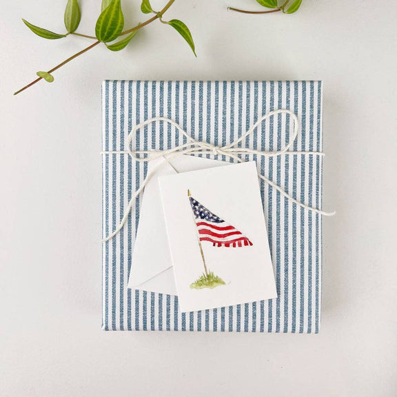 White mini notecard with illustrated American flag, gold pole, and grass, on striped envelope with twine and greenery
