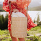 Beige handwoven rush tote with leather shoulder strap, held by person in red patterned dress outdoors