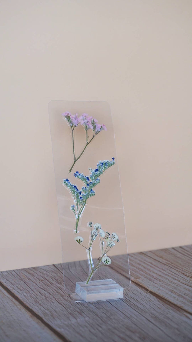 Transparent floral bookmark with pressed purple baby's breath, blue forget-me-nots, and delicate flowers on clear acrylic, white base.