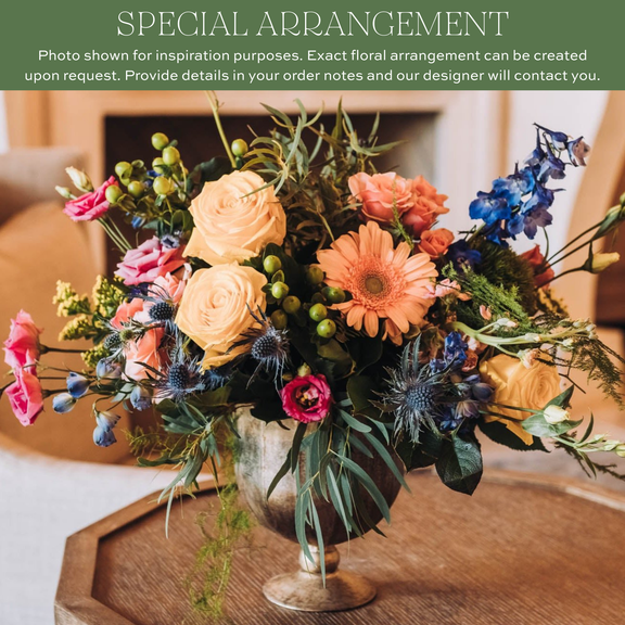 Mixed floral arrangement with peach roses, coral gerbera daisies, pink carnations, blue delphinium, thistle, and green hypericum berries in a clear glass vase