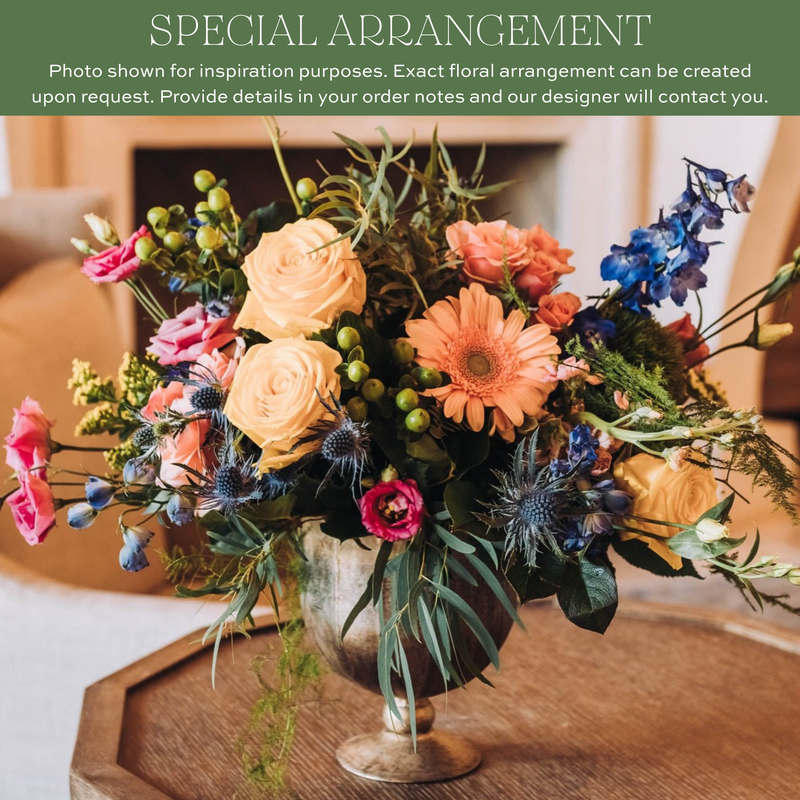 Mixed floral arrangement with peach roses, coral gerbera daisies, pink carnations, blue delphinium, thistle, and green hypericum berries in a clear glass vase