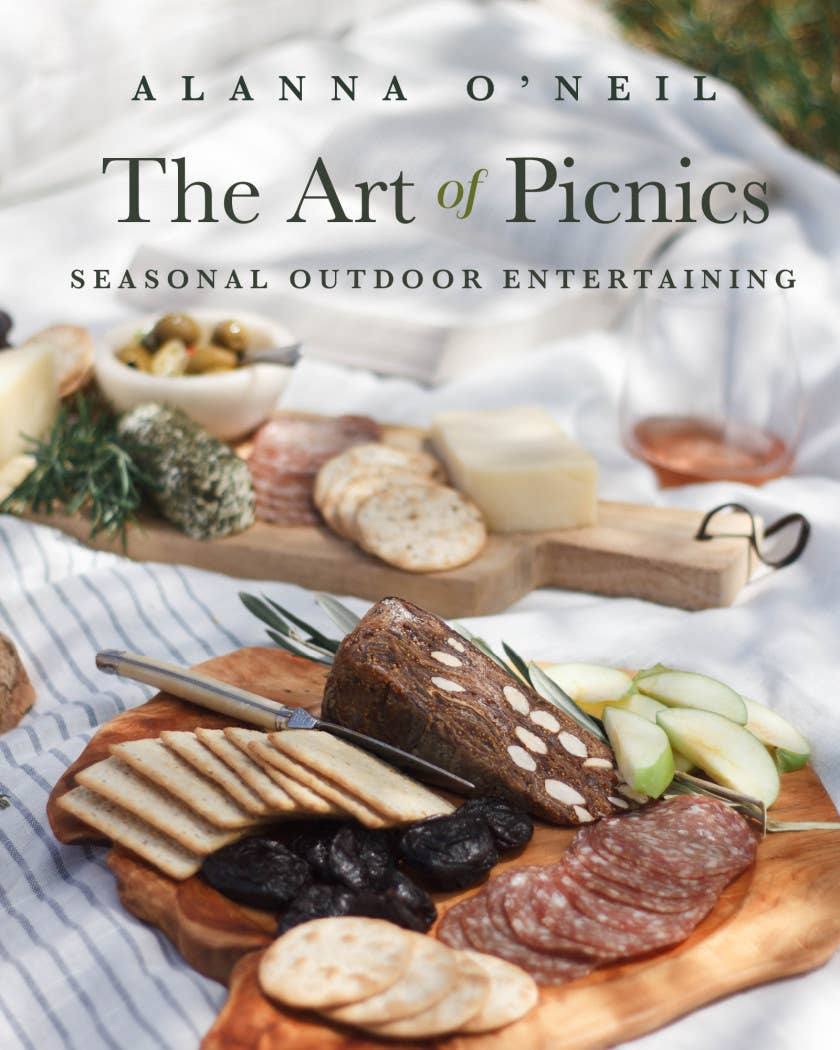 "The Art of Picnics" cookbook cover with author Alanna O'Neill, featuring styled charcuterie boards with bread, cured meats, cheese, and fresh herbs