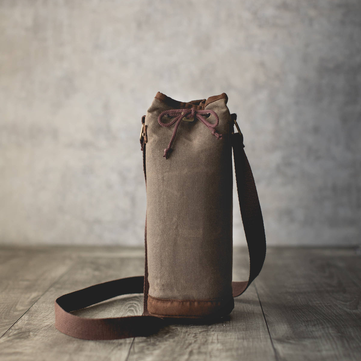 Distressed waxed canvas wine tote with tan exterior, brown leather base, drawstring closure, and black shoulder strap
