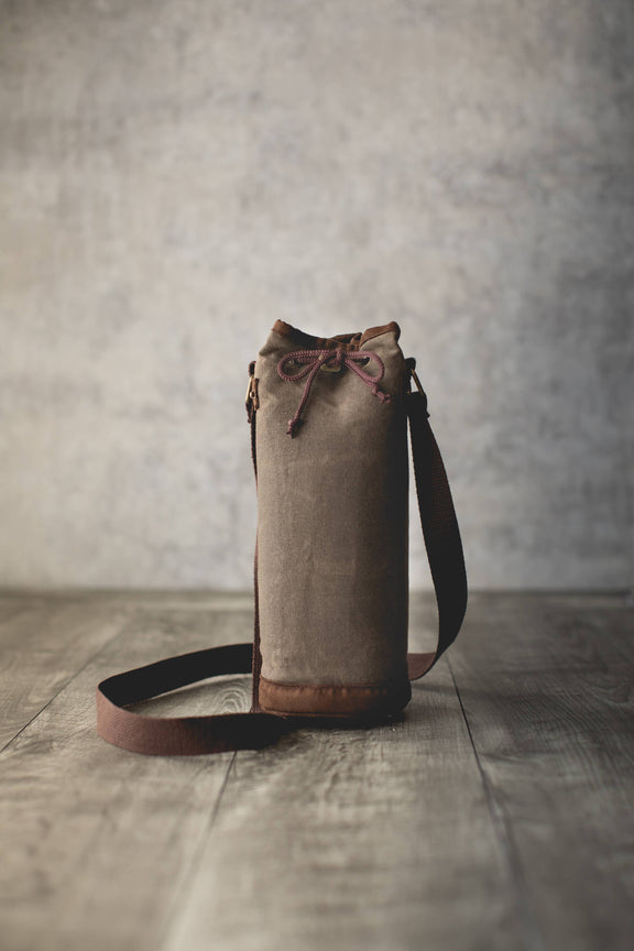 Distressed waxed canvas wine tote with tan exterior, brown leather base, drawstring closure, and black shoulder strap