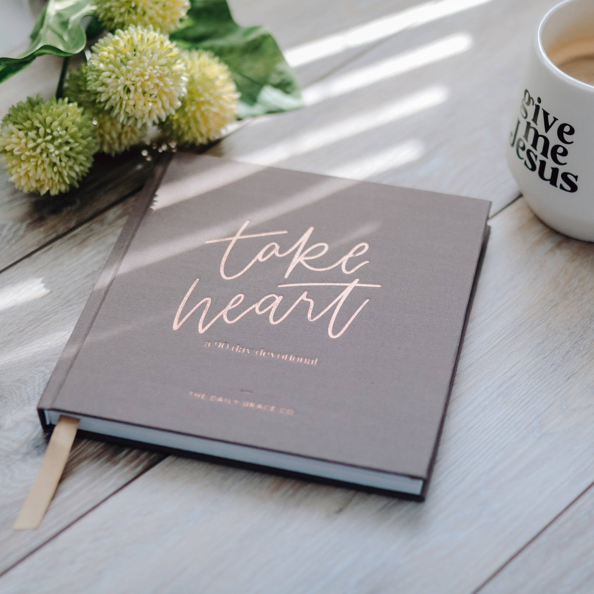 Take Heart devotional book with rose-gold lettering on taupe cover, styled with yellow button mums and white mug on light wood surface.
