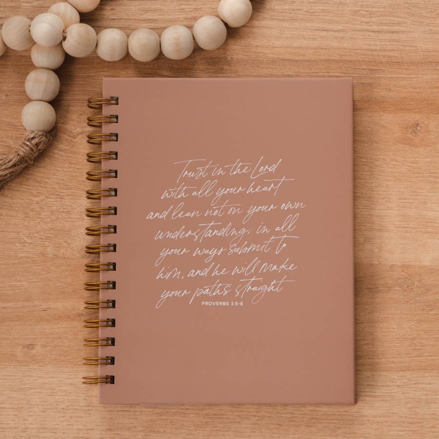 Dusty rose hardcover spiral journal with Proverbs scripture in cursive lettering, styled on wood with wooden beads and twine