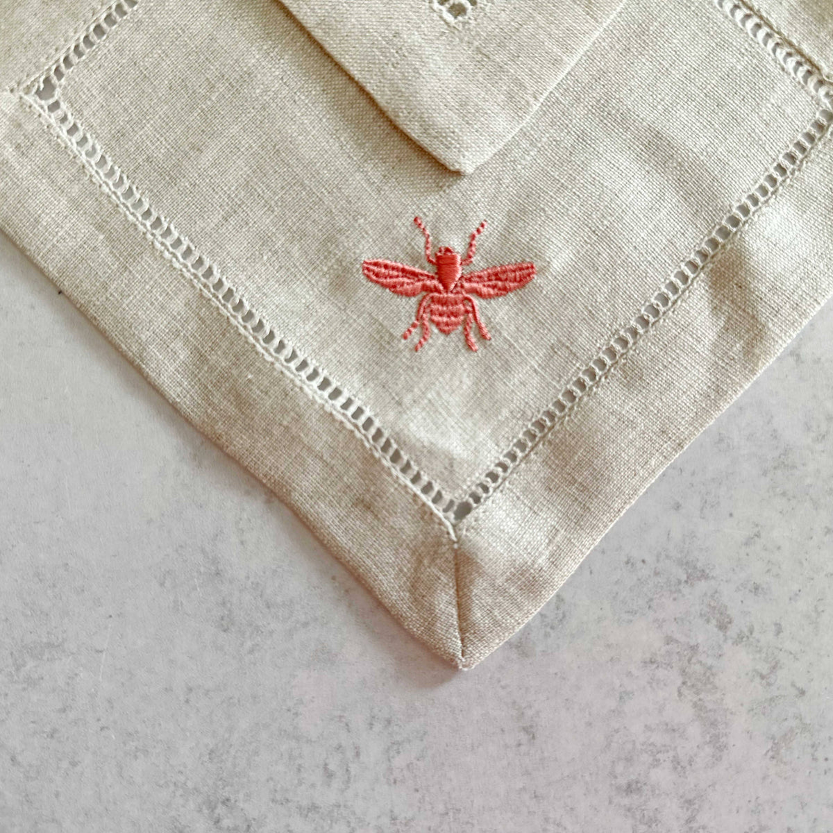 Cream linen square coasters with hemstitched borders and coral-red embroidered bee design