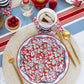 Red floral wave paper plate with white botanical print, set on blue striped tablecloth with striped napkins, fresh raspberries, and gold utensils.