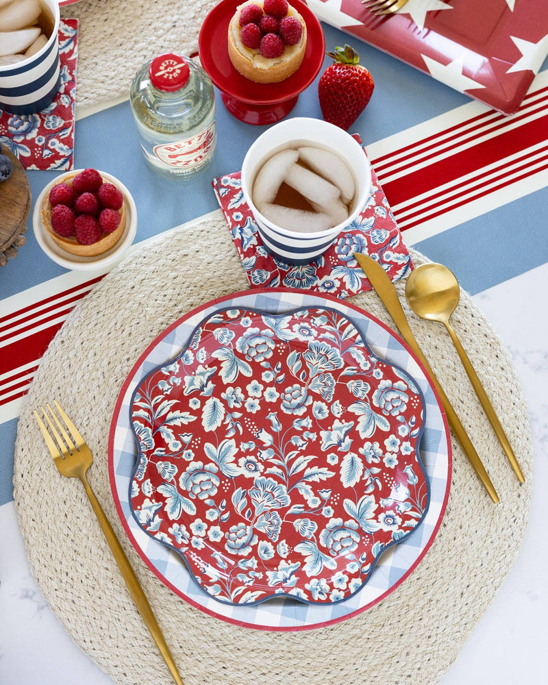 Red floral wave paper plate with white botanical print, set on blue striped tablecloth with striped napkins, fresh raspberries, and gold utensils.