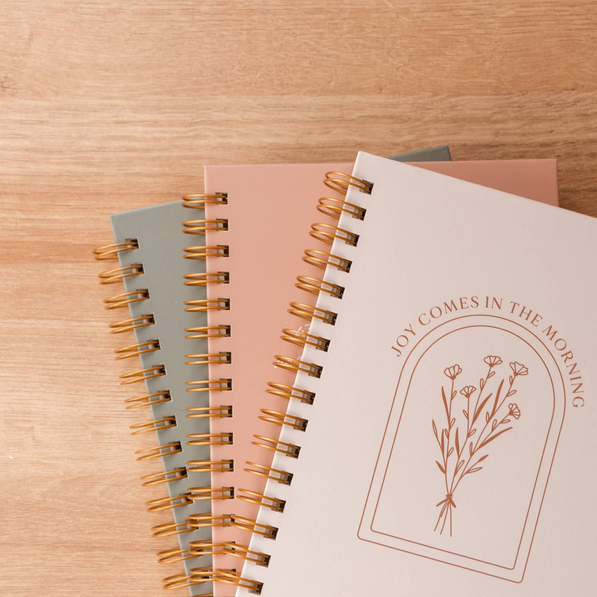 Three hardcover spiral journals with cream, blush and sage covers, gold metal bindings, and botanical flower design on wood surface