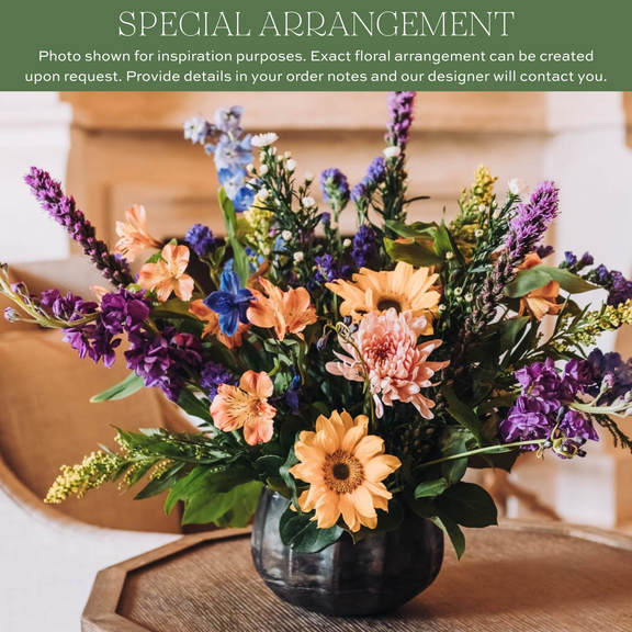 Mixed fresh flower arrangement with peach dahlias, blue delphiniums, purple liatris, and greenery in a dark ceramic vase