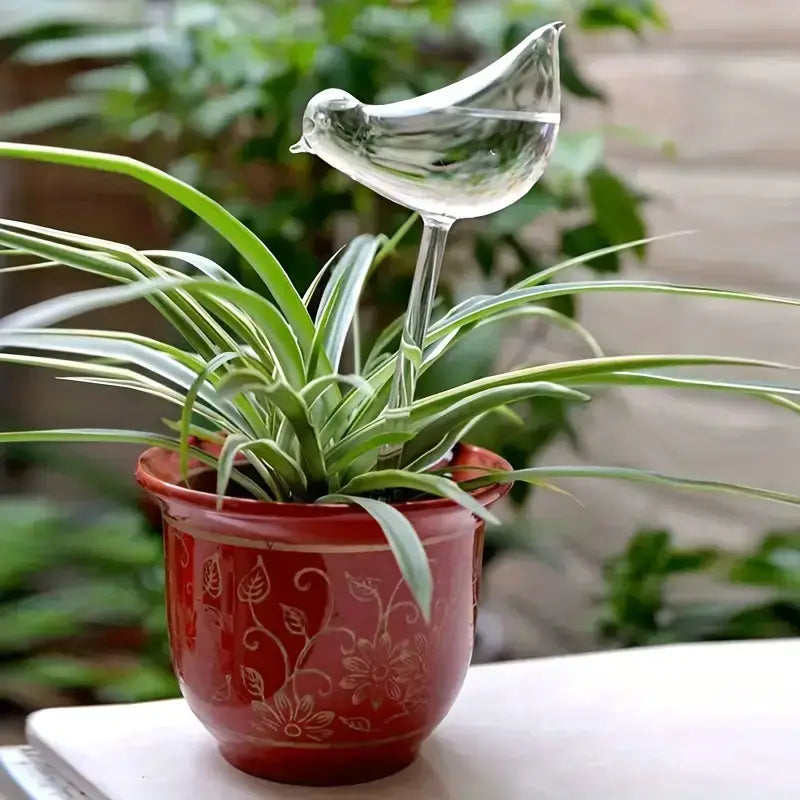 Clear glass bird-shaped watering stake positioned in green potted plant with narrow leaves in decorative red ceramic pot