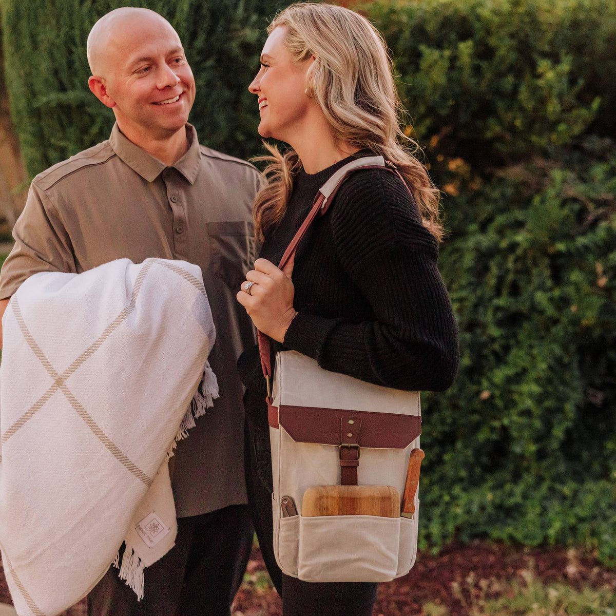 Couple in garden with insulated beige wine tote bag featuring burgundy strap; woman carries wine bottle and cheese board accessories.
