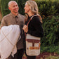 Couple in garden with insulated beige wine tote bag featuring burgundy strap; woman carries wine bottle and cheese board accessories.
