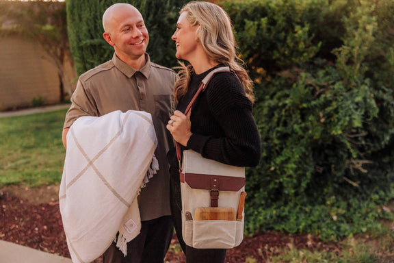 Couple in garden with insulated beige wine tote bag featuring burgundy strap; woman carries wine bottle and cheese board accessories.