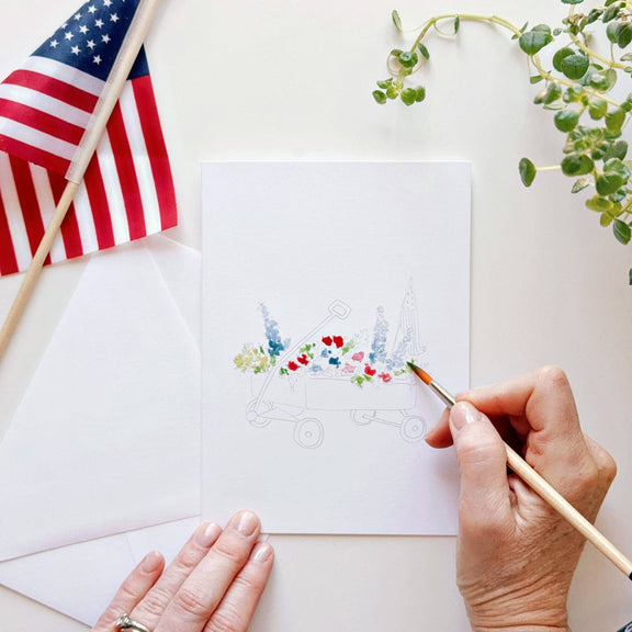 Hand painting a white notecard with patriotic-themed outlines, adding red, blue, and green watercolors to a decorative Fourth of July illustration
