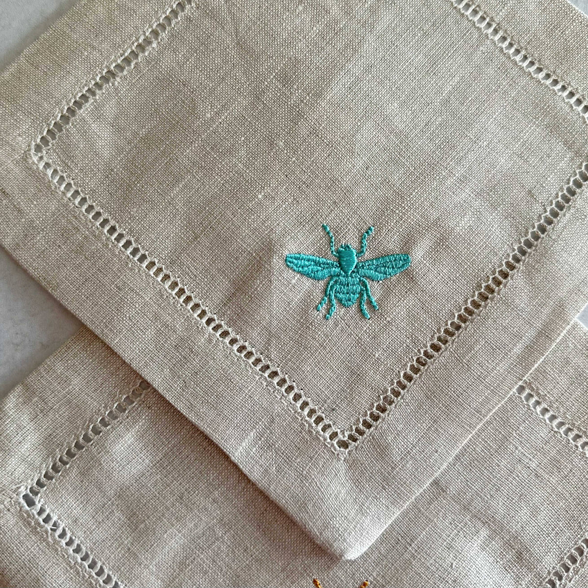 Two stacked natural linen coasters with white hemstitching borders, each featuring a turquoise embroidered bee design