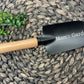 Black garden trowel with wood handle and white engraved "Mom's Garden" text, displayed on woven mat with green succulents