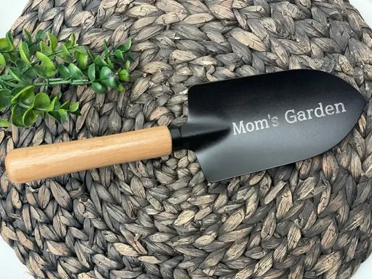 Black garden trowel with wood handle and white engraved "Mom's Garden" text, displayed on woven mat with green succulents