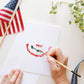 Hand holding paintbrush adding watercolor details to a blank notecard featuring painted red and white firework fan design with blue accents.