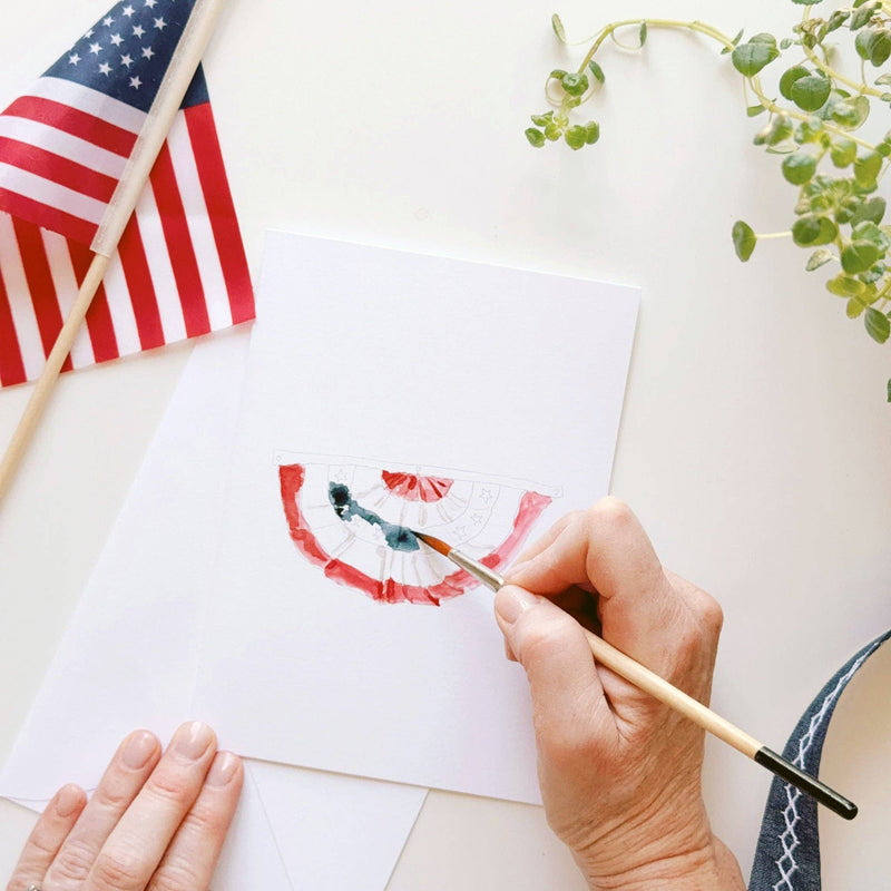 Hand holding paintbrush adding watercolor details to a blank notecard featuring painted red and white firework fan design with blue accents.