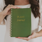 Spiral-bound plant journal with forest green cover and gold lettering, held by hands against a white background