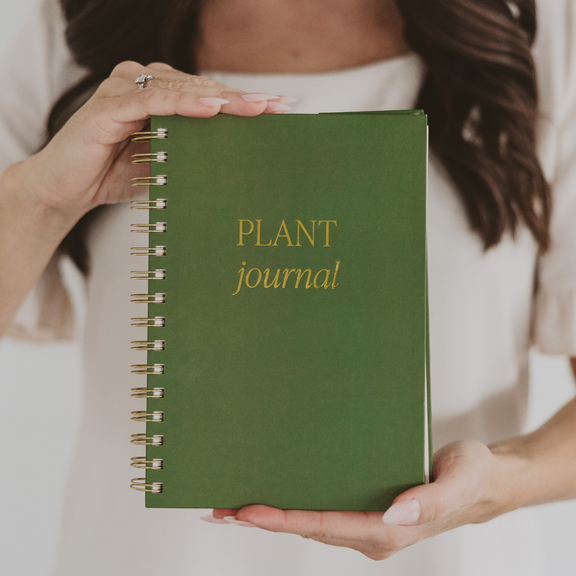 Spiral-bound plant journal with forest green cover and gold lettering, held by hands against a white background
