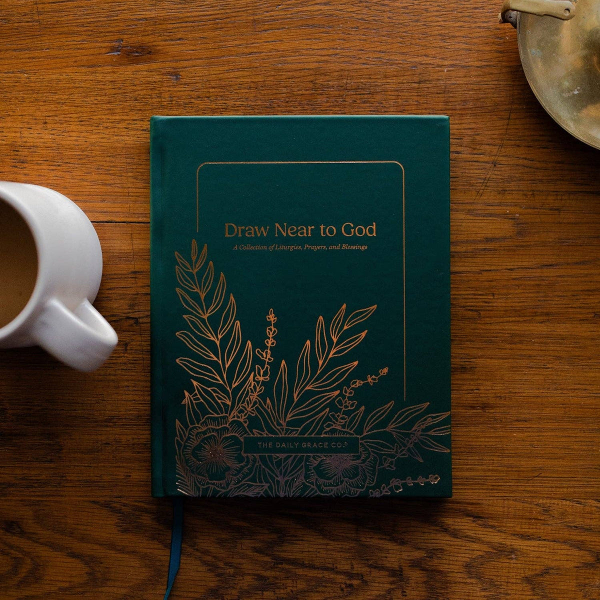 Teal hardcover book titled Draw Near to God with gold foil botanical designs on cover, displayed on wooden surface with coffee mug.